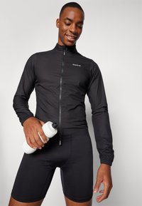 Svart cykeljacka med hög krage och dragkedja fram, kombinerad med svarta shorts. Hand som håller en vit vattenflaska.