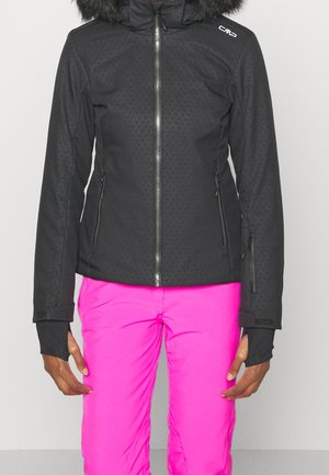 Woman wearing a black zip-up winter jacket with fur-trimmed hood and bright pink snow pants, standing against a plain background.