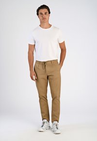 Weißes T-Shirt mit kurzen Ärmeln, hellbraune tapered Hose mit Zugband und weiße Sneakers mit bunten Akzenten, vor einem einfarbigen Hintergrund stehend.