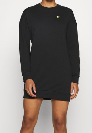 Femme portant une robe sweat-shirt noire à manches longues avec un petit logo d'oiseau jaune sur la poitrine, debout devant un fond uni.