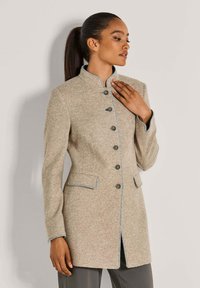 Vrouw in een beige button-up jas met een staande kraag en contrasterende afwerking, poseert met één hand bij haar borst tegen een effen achtergrond.