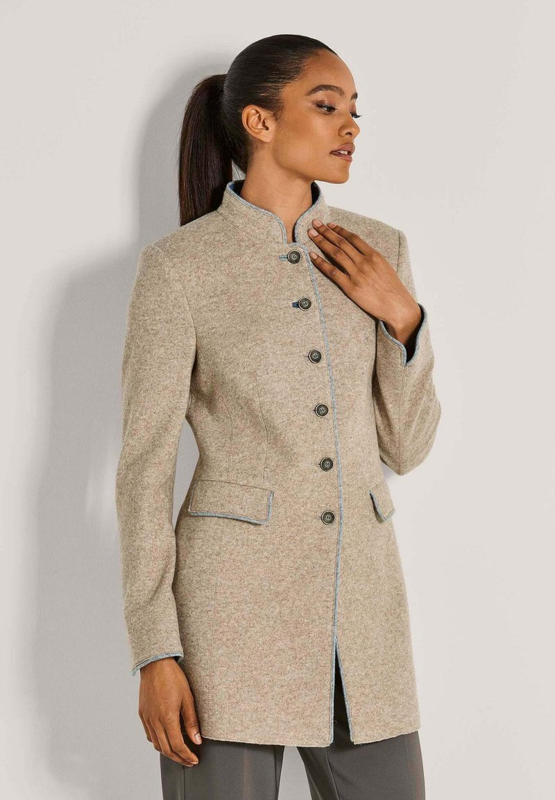 Vrouw in een beige button-up jas met een staande kraag en contrasterende afwerking, poseert met één hand bij haar borst tegen een effen achtergrond.