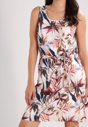 Woman wearing sleeveless, knee-length dress with colorful tropical leaf pattern and fabric belt tied at the waist on light background.