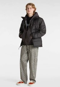 Czarna puchowa kurtka z kapturem nałożona na ciemnoszarą bluzę z kapturem. Do tego luźne, wzorzyste zielone spodnie i brązowe trampki.