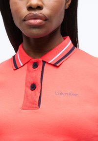 Γυναίκα που φοράει κοραλί πόλο μπλουζάκι Calvin Klein με γιακά σε ναυτικό μπλε και λευκές ρίγες και δύο μαύρα κουμπιά κουμπωμένα στον λαιμό.