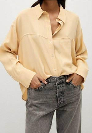 Vrouw draagt een loszittend, lichtgeel overhemd met knopen, ingestopt in een high-waisted, vervaagde grijze jeans met handen in de zakken.