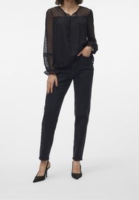 Blouse noire transparente à pois, col noué et manches bouffantes, associée à un jean skinny noir et des escarpins noirs pointus.
