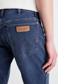 Vaqueros de mezclilla con un lavado de azul oscuro, que cuentan con un parche de cuero de Wrangler en el bolsillo trasero y costuras naranjas en un patrón de W.