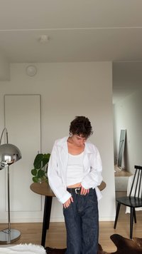 Camicia bianca oversize con bottoni, canotta bianca aderente, jeans larghi in denim scuro, cintura nera con fibbia argentata, ambientazione interna con pianta.