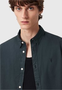 Camicia button-up verde scuro in tessuto leggero con colletto, caratterizzata da bottoni bianchi e un sottile logo ricamato sul petto.