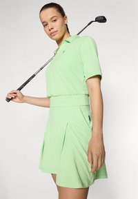 Vestido de golf verde claro con mangas cortas, falda plegada, textura acanalada y una cinturilla ajustada, complementado con un palo de golf en la mano.
