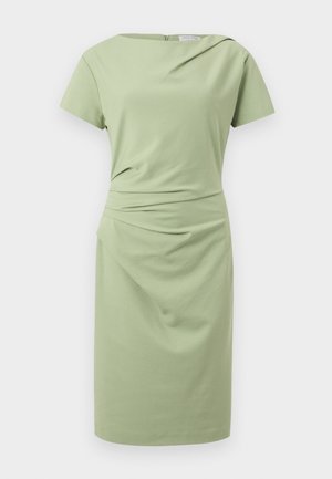 Kvinde i lysegrøn knælængde kjole med korte ærmer, beige spidse sko og sort skuldertaske, stående med hånden på hoften.