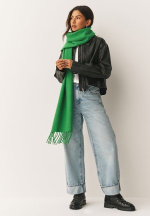 Donna con giacca di pelle nera, jeans azzurri a gamba larga, stivali neri e lunga sciarpa verde in piedi contro uno sfondo semplice.