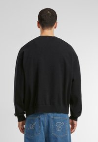 Sudadera negra de cuello redondo con hombros caídos, puños y dobladillo acanalados, combinada con jeans azules que tienen costuras decorativas en los bolsillos.