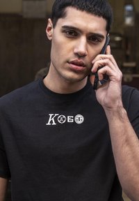 Musta lyhythihainen t-paita, jossa on valkoiset logot "K", "KG" ja "G" rinnassa. Paita on valmistettu sileästä puuvillasta ja siinä on pyöreä pääntie.