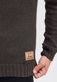 Maglione lavorato a maglia verde scuro con polsini e orlo a costine. Presenta una toppa in pelle con logo rettangolare sul lato inferiore sinistro.