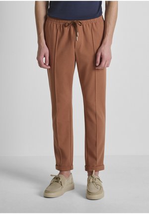 Homme portant un pantalon marron à cordon avec des ourlets retroussés et des chaussures beige à lacets, debout devant un fond gris clair uni.