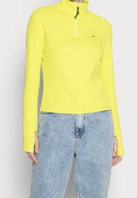 Yellow, ribbed long-sleeve top with a half-zip collar and thumbholes. Paired with light blue, high-waisted jeans.