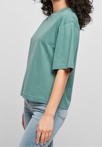 Femme portant une chemise ample à manches courtes bleu sarcelle et un jean bleu clair, debout de profil devant un fond clair uni.