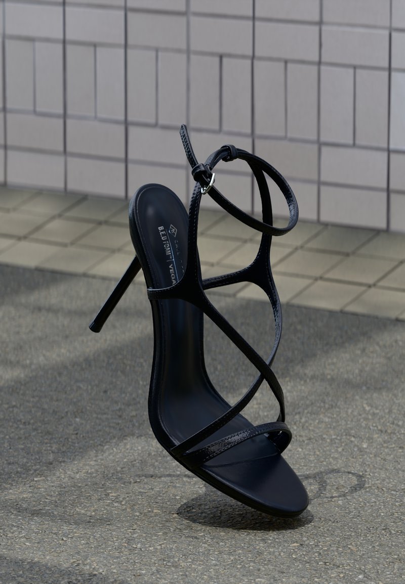 Μαύρο σανδάλι με ψηλό τακούνι, λεπτά λουράκια και αγκράφα στον αστράγαλο, τοποθετημένο όρθιο σε γκρι πλακόστρωτο κοντά σε πλακιδωτό τοίχο.