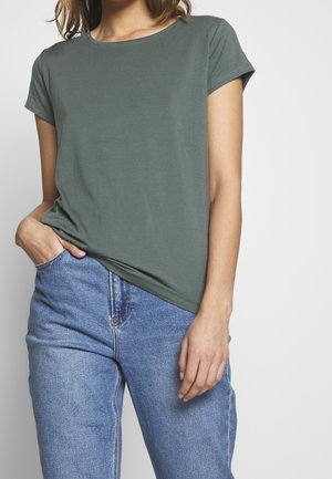 Vrouw met een olijfgroene T-shirt met korte mouwen, lichtjes ingestopt in een hooggetailleerde blauwe spijkerbroek, staand tegen een effen lichte achtergrond.