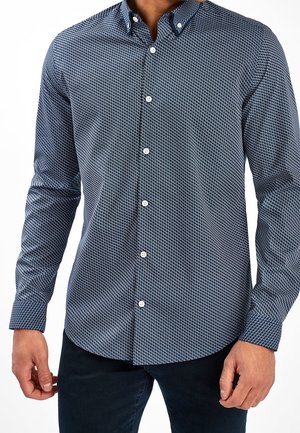 Homme portant une chemise bleu marine à manches longues avec un petit motif géométrique blanc et un pantalon foncé.