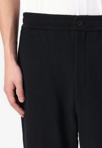 Mustad ribadega mustad püksid laia elastse vöökohtaga, millel on nuppluku, ja siledaks tekstuuriga, osaliselt näidatud käega, mis puhkab nende peal.