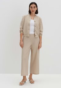 Beige Zip-Jacke mit Rundhalsausschnitt und goldenen Knöpfen, kombiniert mit hoch taillierten, weiten Hosen und beigen Sandalen. Glatte Stofftextur.