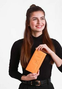Oranžová peněženka s hladkou texturou, obdélníkovým tvarem a decentním designovým detailem uprostřed. Držena v lidských rukou na jednobarevném pozadí.