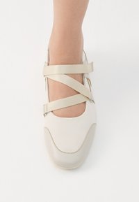 Lys beige ballet flat med kryssremmer. Har en glatt stoffoverdel og en litt mørkere, blank tåhette. Enkel og stilren design.