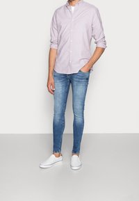 Hombre vestido con una camisa de botones de color púrpura claro, jeans ajustados azules, zapatos de lona blancos, de pie con una mano en el bolsillo contra una pared lisa.