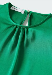 Blouse verte avec encolure ronde, ouverture goutte d'eau au dos et petite fermeture par bouton, fabriquée en tissu lisse.