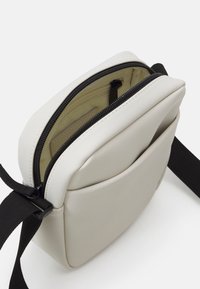 Borsa a tracolla compatta di un beige chiaro con spallaccio regolabile nero, tasca frontale e scomparto principale parzialmente aperto che mostra la fodera in tessuto beige e una tasca interna con zip.