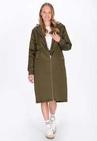 Hosszú, olíva zöld párnázott kabát, elöl cipzárral, szürke kapucnival, oldalsó zsebekkel és bordázott mandzsettával, fehér ing fölött viselve, sportcipővel párosítva.