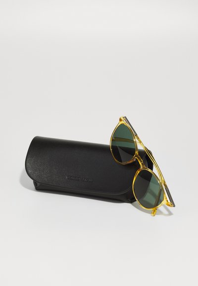 Ochelari de soare cu ramă galbenă și lentile verzi, așezați pe un toc din piele neagră cu un logo embosat. Textură netedă și design rotunjit.