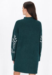 Türkis gestricktes Kleid mit hohem Kragen und langen Ärmeln, verziert mit silbernen, paillettenbesetzten Blumenmotiven auf den Ärmeln. Weiche Textur.