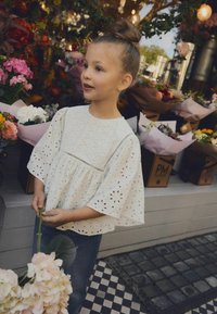 Blouse blanche en broderie anglaise avec des manches larges, ornée de découpes circulaires ; portée avec un jean bleu. Un bouquet de fleurs roses claires est tenu.