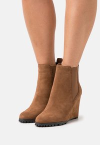 Botins de tornozelo em camurça castanha com painéis laterais elásticos e solas texturizadas. Acabamento suave, bico arredondado e uma alça na parte de trás.