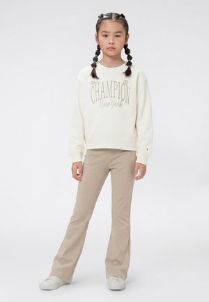 Jeune fille aux cheveux tressés portant un sweat-shirt Champion crème, un pantalon beige et des baskets blanches, debout devant un fond uni.