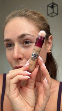 Multi-Anwender Concealer von Maybelline in einem transparenten Röhrchen mit bordeauxrotem Deckel, der ein beige farbenes Produkt zeigt. Eine Frau hält ihn nahe an ihr Gesicht.