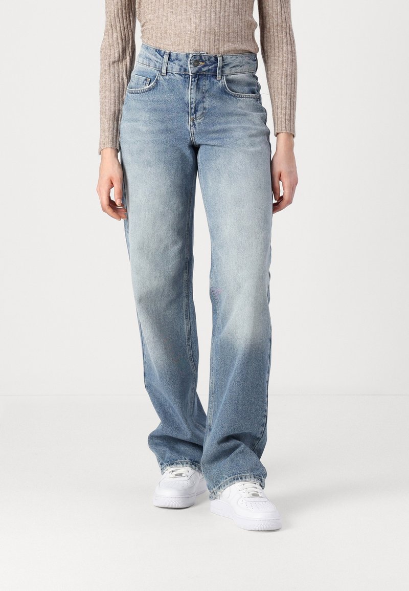 Personne portant un jean large bleu clair, un haut beige à manches longues côtelé, et des baskets blanches, debout devant un fond blanc uni.