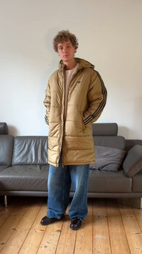 Dun, puffy vinterjakke med hette, svarte striper på ermene, lommer, båret over en lys skjorte, kombinert med vide blå jeans og mørke sko.