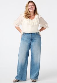 Roomwitte blouse met ruches en driekwart mouwen, gecombineerd met lichtblauwe jeans met wijde pijpen. Draagt schoenen met een patroon en een textuurafwerking.