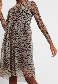 Leopardenmusterkleid mit langen durchsichtigen Ärmeln, fließender Silhouette, runder Ausschnitt und geraffter Taille, in Beige- und Schwarztönen.