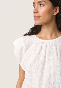 Vit broderad blus med korta volangärmar, prydd med blommönster, med en samlad halsringning och lätt, texturerad tyg.
