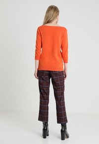 Pull en tricot orange à manches trois quarts, associé à un pantalon à carreaux marron, avec une taille haute et un bas évasé, et des bottes à talons noires.