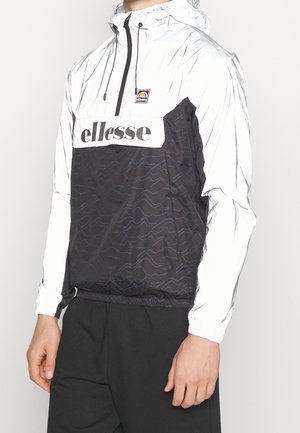 Osoba ubrana w czarno-białą kurtkę z kapturem ellesse typu wiatrówka z przednim zamkiem błyskawicznym i logo na piersi, w połączeniu z czarnymi spodniami.