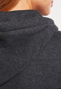 Šedá mikina s kapucí představující detailní pohled na strukturovanou tkaninu, vybavená stahovací šňůrkou na kapuci a hladkým povrchem s jemnými odstíny.