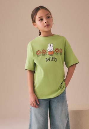 Mädchen mit geflochtenem Haar trägt grünes Miffy-T-Shirt mit orangefarbenen Blumen, kombiniert mit locker sitzenden hellblauen Jeans, steht vor einer schlichten beige Wand.