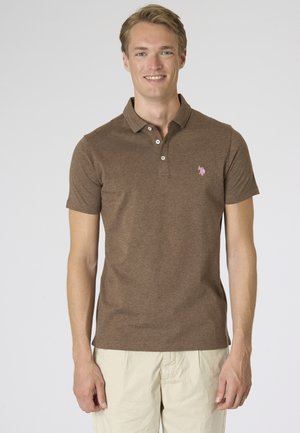 Giovane uomo sorridente, indossa una polo marrone a maniche corte e pantaloni beige chiaro, in piedi davanti a uno sfondo semplice.